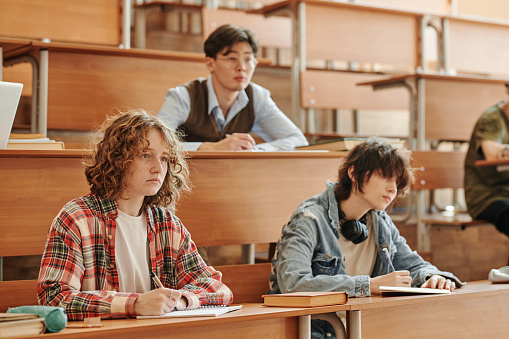 Group of youthful interracial students in casualwear making notes in copybooks while sitting by long desks in lecture hall
