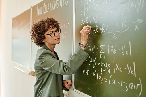 Serious female teacher of algebra with piece of chalk writing down equations on blackboard while explaining new subject