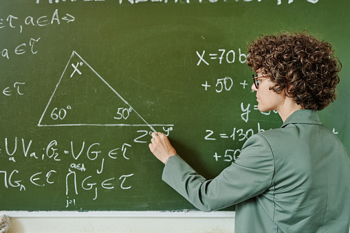 Young serious teacher of college or university drwing triangle on blackboard while explaining theorem to audience