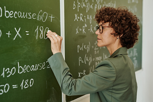 Side view of young teacher with short dark curly hair writing down equation on blackboard while explaining it to students
