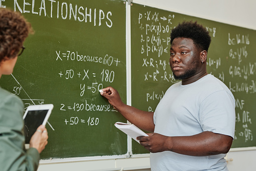 Youthful African American student pointing at blackboard during explanation of equation or theorem and looking at teacher