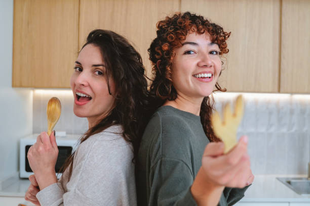 Happy Latin female friends having fun dancing while cooking together at home kitchen - Focus on right girl face