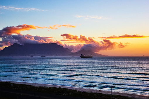 Clouds over the South African landmark of Table Mountain light up at sunset in shades of yellow, orange and purple, with the sea of Table Bay in the foreground.