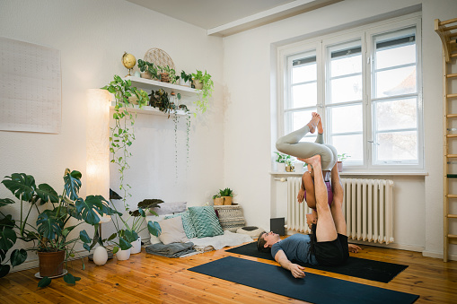 Young couple practising acro yoga poses at home. Man lying on yoga mat and balancing a woman upside down on his legs while exercising at home.