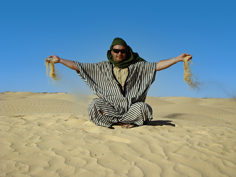 A man in traditional Berber costume sits on a dune in the Sahara desert, Tunisia. Sand spills from his palms. Close-up.