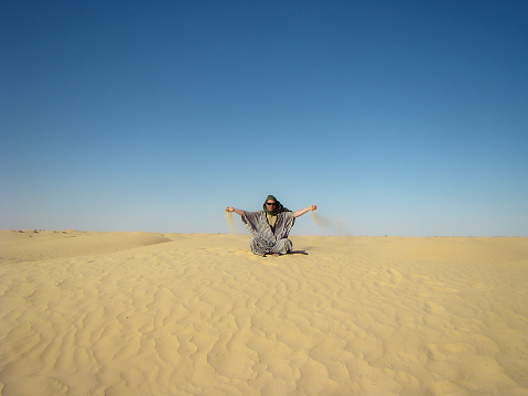 A man in traditional Berber costume sits on a dune in the Sahara desert, Tunisia. Sand spills from his palms.