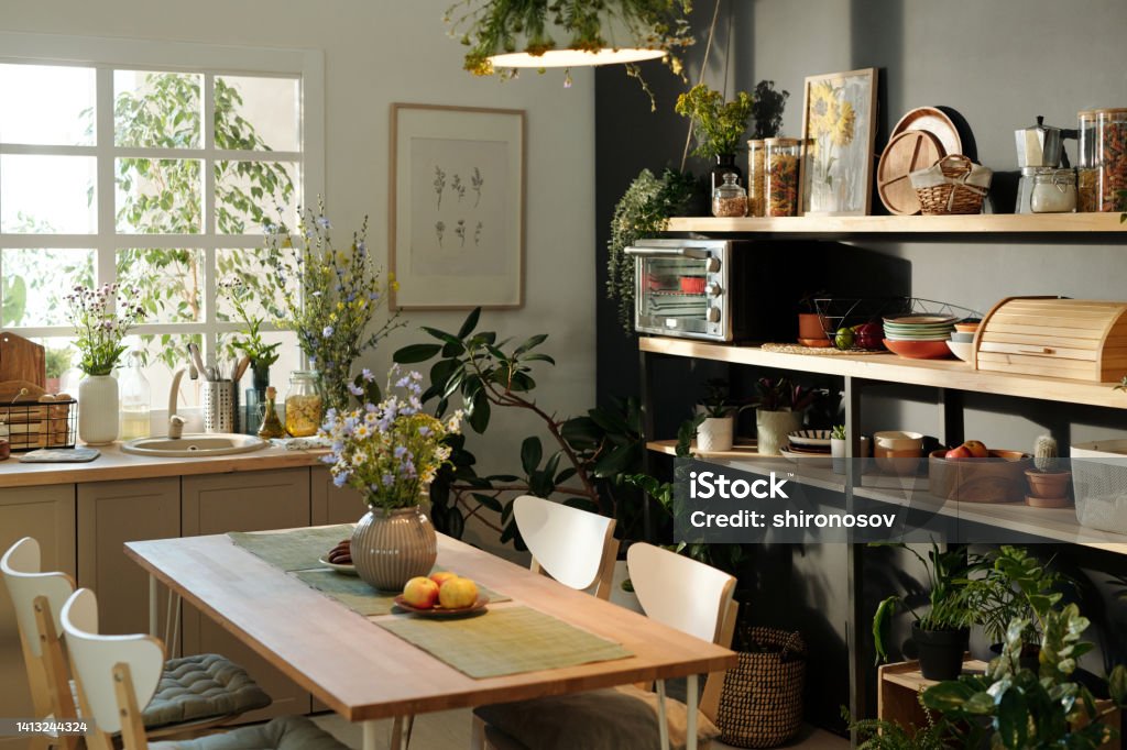 Interior of cozy kitchen with table and chairs in the center against window Interior of cozy kitchen with chairs surrounding table with napkins, wildflowers in vase and apples on plate standing by window Apartment Stock Photo