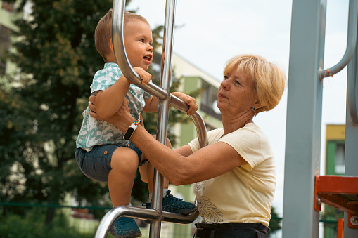 Grandmother supporting and holding her 2 years old grandson at children playground. Grandmother and child having fun. Support childhood symbol. Sunny, summer day.