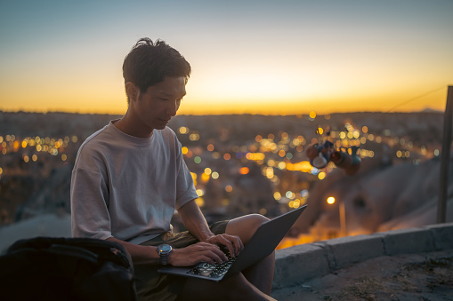 A young male freelance photographer videographer tourist is using a laptop on a hill during sunset during his vacation.