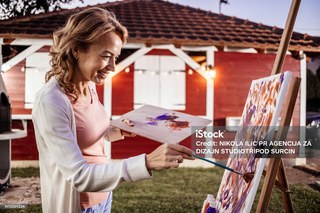 Female artist painting at home stock photo Portrait of blonde woman painting for hobby in her back yard studio, practicing with paint, brushes, easel, canvas and colours, wearing white sweater and pink shirt Painted Image Stock Photo