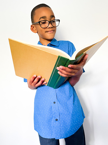 Young black male wearing a blue shirt, dark jeans, and black glasses. He has a inquisitive expression while holding and reading a big textbook