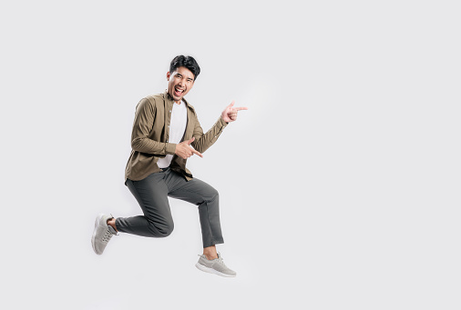 Full length excited handsome young Asian man jumping in air with hand pointing to empty space on white background. Cool man joyful running in copy space. Studio short.