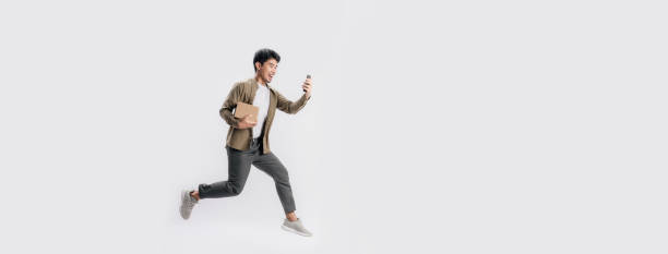 Full length handsome Asian man holding box and use smartphone he is run and jumping in air on banner white background. stock photo