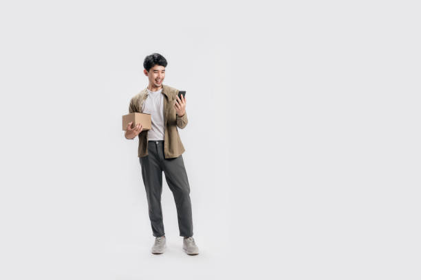 フルレングスのハンサムなアジアの男性は、ボックスを保持し、孤立した白い背景にスマートフォンを使用しています。 - asia businessman asian ethnicity happiness ストックフォトと画像