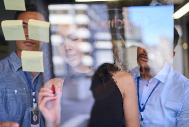 planning, training and presenting businesswoman writing sticky notes and strategizing a project on a glass wall in an office. group of colleagues brainstorming an idea or strategy in a startup agency - cheering business three people teamwork imagens e fotografias de stock