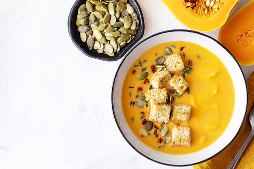 Autumn pumpkin cream soup in bowl served with seeds and crouton on white stone table top view with copy space.