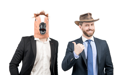 Happy man point finger at weird businessman wearing horse head mask in formalwear isolated on white, western party.
