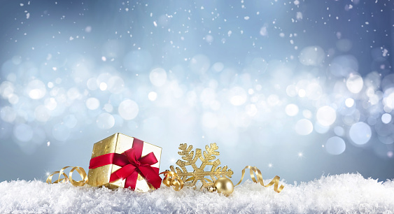 Festive Christmas snow background with copy space. Golden gift box with red ribbon, snowflake and serpentine on snow on blue background with beautiful bokeh circles.