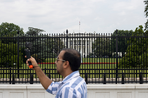 Washington, DC, USA - June 22, 2022: A tourist is seen recording a video with his action camera in the blurry foreground outside the White House in Washington, DC.