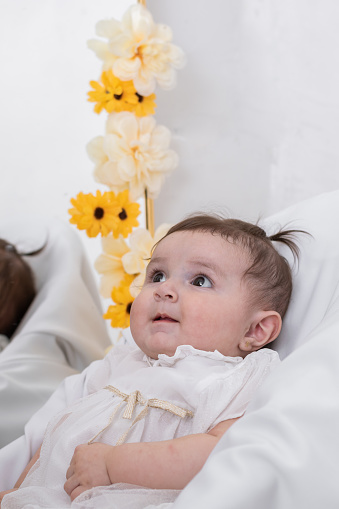 beautiful latin baby girl, very happy looking at her mother, lying on a white sheet next to a mirror. six month old baby girl in a white dress.