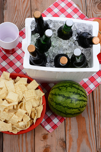 High angle shot of a picnic spread on a wood deck with red and white checkered table cloth. Items include: beer, ice chest, cups, chips and watermelon