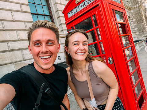 Mobile Phone Handheld Selfie of Two Caucasian Male and Female Tourist Traveler Friends Smiling Together in Front of an Iconic Red Telephone Booth in London, United Kingdom