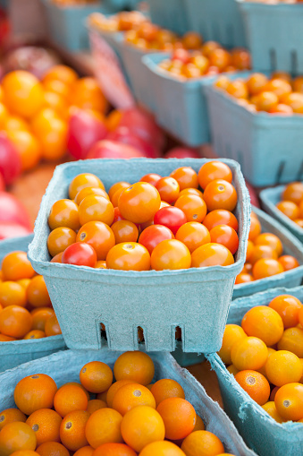 Close-up of colorful yellow and red cherry tomatoes, in front of assorted heirloom tomatoes, displayed in stacked blue cartons at a farmer's market