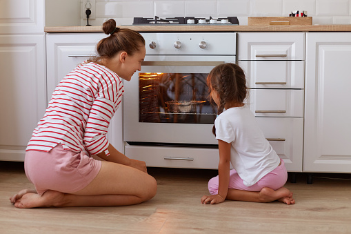 istock Portrait of little dark haired kid watching with her mother at oven, baking cookies in oven indoors, family wearing casual clothing, sitting on floor in kitchen. 1413300459