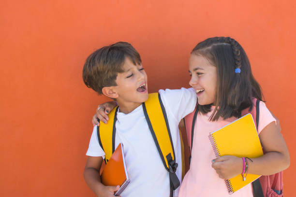 Elementary age children with facial expression of joy on the first day of school. Elementary age children with facial expression of joy on the first day of school. Smiling preschool girl and boy of 7s with backpacks and notebook with orange background.Children with funny expression back to school stock pictures, royalty-free photos & images