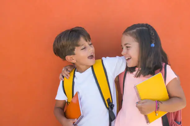 Elementary age children with facial expression of joy on the first day of school. Smiling preschool girl and boy of 7s with backpacks and notebook with orange background.Children with funny expression