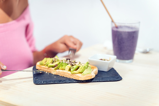 Close-up view of a woman eating an avocado toast and drinking smoothie while having breakfast. Healthy food concept.