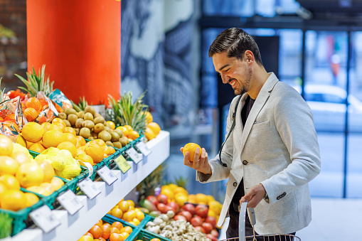 A young Caucasian businessman is standing in the fruit aisle of a supermarket and holding a fresh orange with a wide smile on his face.