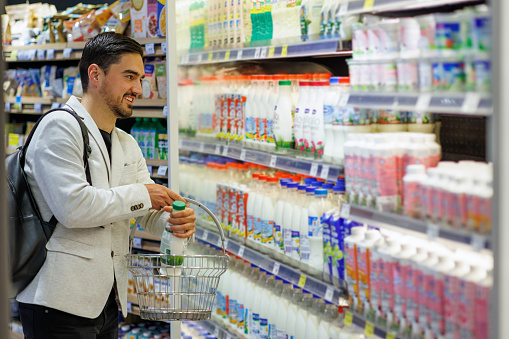 A young Caucasian businessman is standing in front of the dairy products in a supermarket and picking a bottle of milk with a smile on his face.