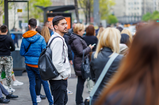 A young Caucasian business person wearing a backpack is standing at a bus stop and patiently waiting for his ride with his hands in his pockets.