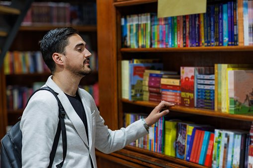A young Caucasian man wearing a backpack is standing  in a bookstore and picking out books from one of the shelves.