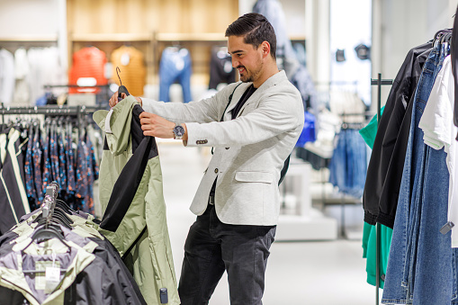 A young Caucasian man is standing in a clothing store and checking out a jacket with a smile on his face.