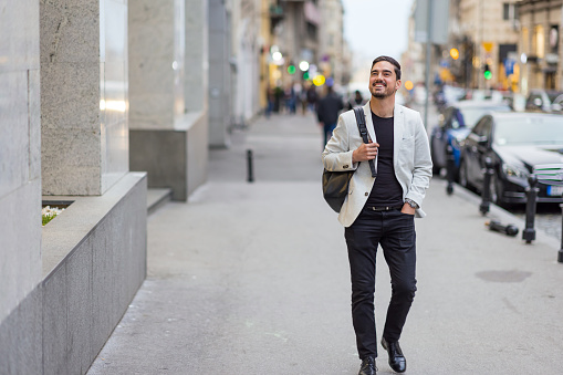 A young Caucasian businessman carrying a backpack is walking down the street and looking around with a smile on his face.