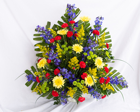Funeral arrangement of yellow cremones, solidaster, red carnations, blue delphinium, kermit poms, ferns. White background
