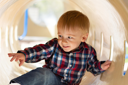 Toddler boy in plaid shirt and jeans scoots through playground tunnel