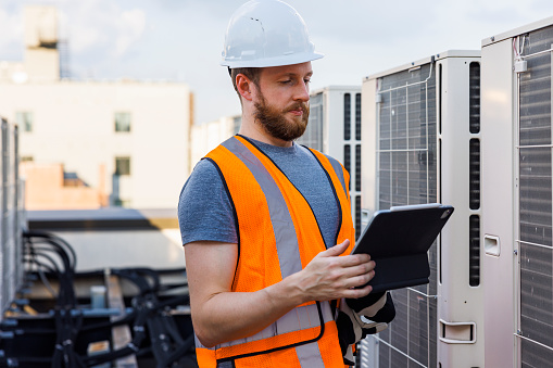 Rooftop air conditioning system inspection. Caucasian white workers check units and log the result with a portable computer.