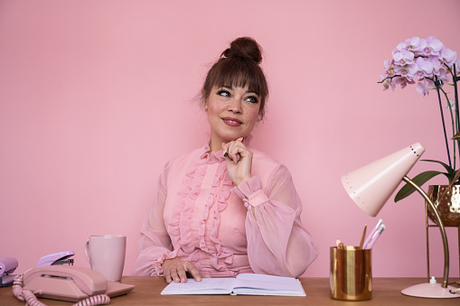 Waist up front view of well-dressed mature woman working amidst retro style office accessories and looking away from camera against pink background.
