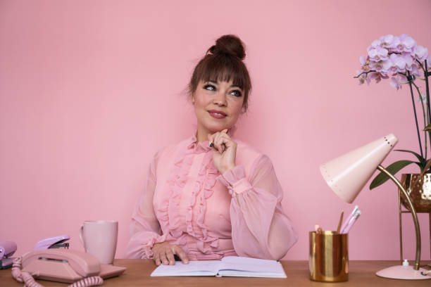 Portrait of businesswoman in ruffled blouse sitting at desk Waist up front view of well-dressed mature woman working amidst retro style office accessories and looking away from camera against pink background. topknot stock pictures, royalty-free photos & images