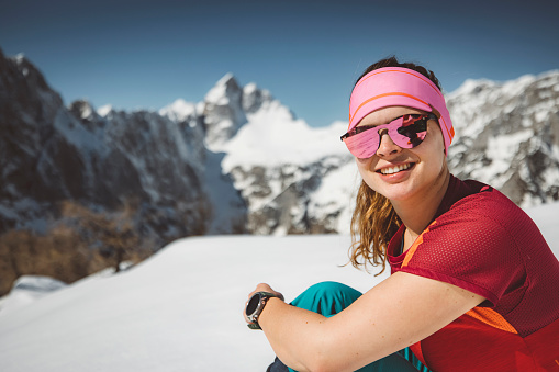 Portrait of smiling caucasian woman hiker on a winter hike, beautiful sunny day outside and a lot of snow in the mountains. Woman wearing a headband and sunglasses.