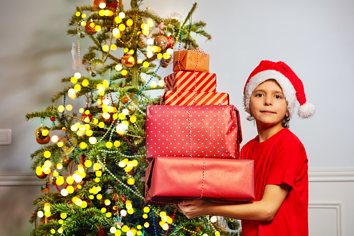 Profile photo of little boy stand with huge stack of presents, in Santa hat near Christmas tree