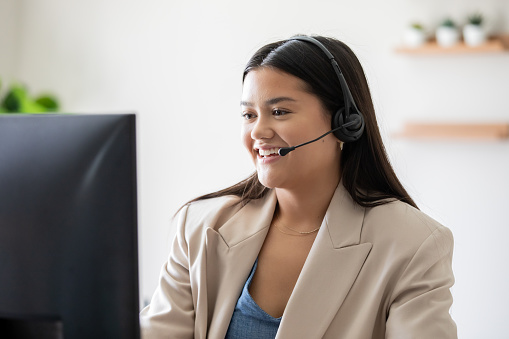 Young woman working in call center takes calls on headset while looking at computer screen