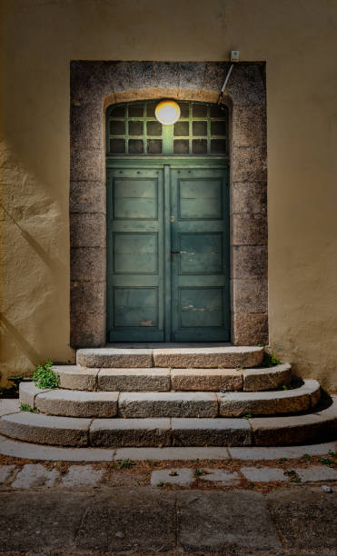 Old double wooden door with glass window at the top, lit by lamp. Yellow plaster outer wall. Stairs to enter the building. Evening scene. Ajaccio, Corsica.