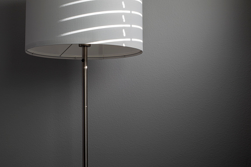 White textile floor lamp with sun light and shadows from window shades. Gray textured wall on the background. Minimalist style.