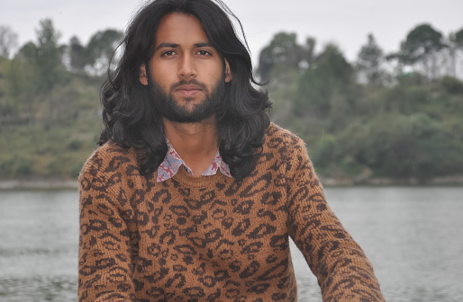 Close-up of a attractive Indian young guy with long hair and beard looking at camera while sitting by the lake