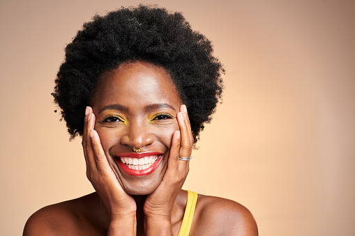 Closeup portrait of a happy, young and beautiful black woman with full glam makeup and a great skincare routine. A dark skinned female with an amazing smile and afro holding her face in joy.
