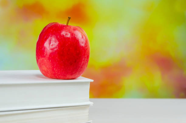 Stack of white books with juicy fresh apple on desktop over blurry autumn background. stacked books with red apple. Background for school design. Back to school.